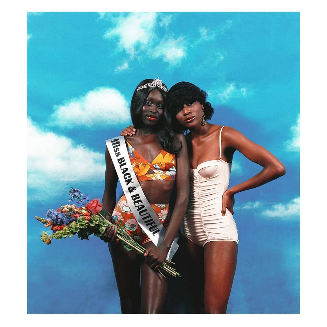Two dark-skinned women in swimsuits pose in front of a cloud-painted backdrop; one holds flowers and wears a beauty pageant sash and crown.