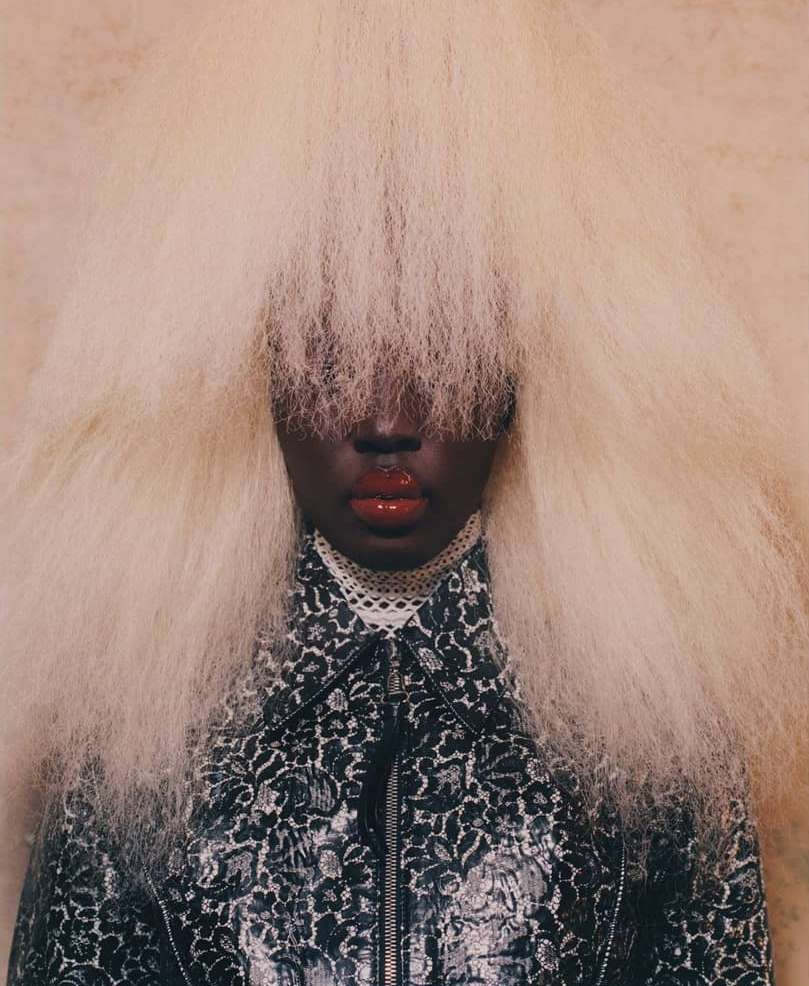 Close-up studio shot of an African American woman with dark skin and red lips wearing a patterned jacket and blonde afro-style wig.