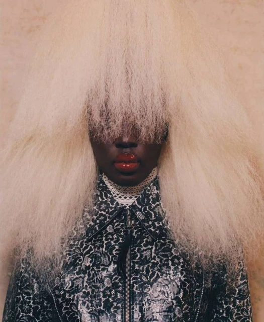 Close-up studio shot of an African American woman with dark skin and red lips wearing a patterned jacket and blonde afro-style wig.