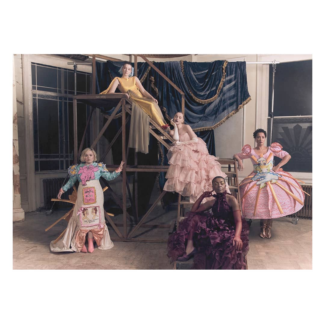 Five women in elaborate dresses pose on and around a metal scaffolding indoors with a draped curtain in the background.
