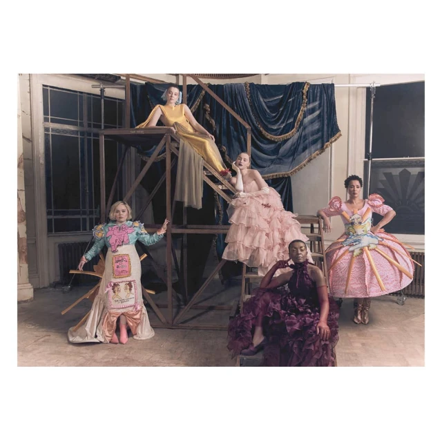 Five women in elaborate dresses pose on and around a metal scaffolding indoors with a draped curtain in the background.