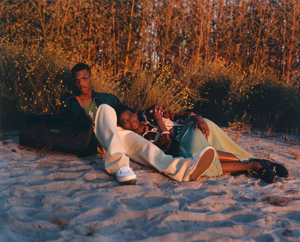 A young couple relaxes on a sandy beach surrounded by tall grass and wildflowers in golden light, with a Gucci bag visible.