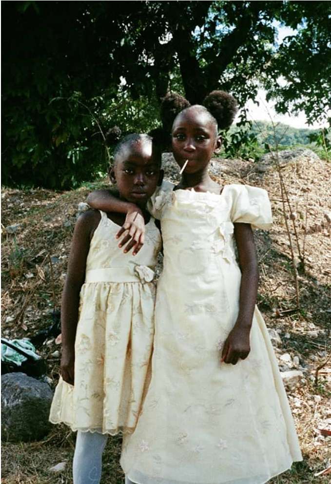 Two young African girls in formal dresses stand together outdoors in front of a tree and vegetation. One girl has a lollipop.