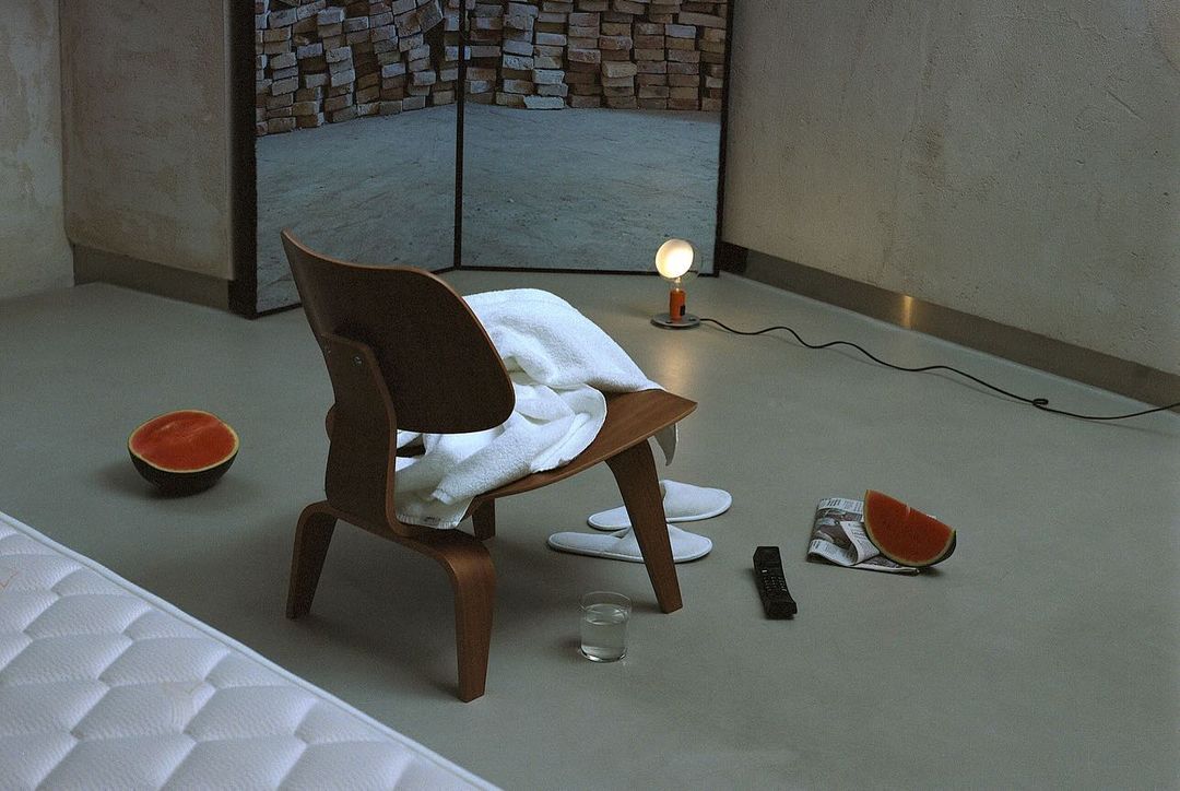 Minimalist interior scene features a wooden chair with a white towel, watermelon slices, slippers, and a small table lamp on a gray floor.