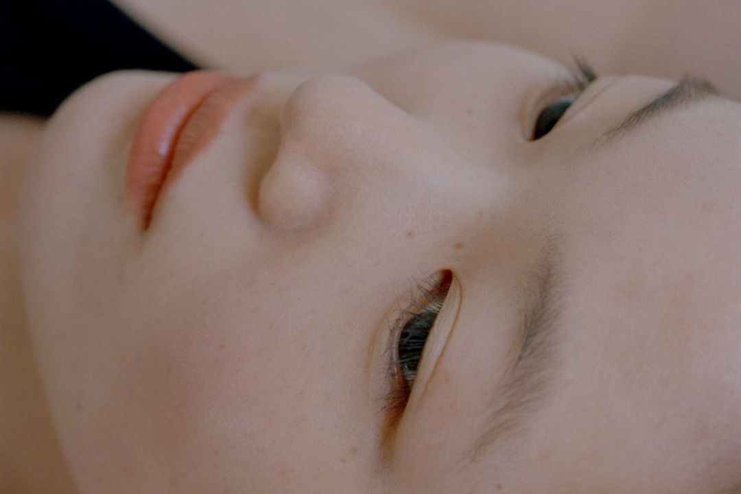 Close-up of a young adult female's face, focusing on her eyes, nose, and lips against a blurred dark background, highlighting makeup.