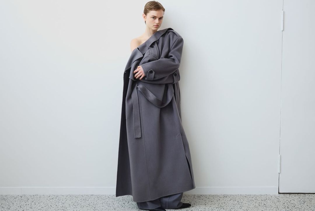 A fair-skinned woman poses indoors wearing a long grey belted coat, looking at the camera in a fashion studio shot.