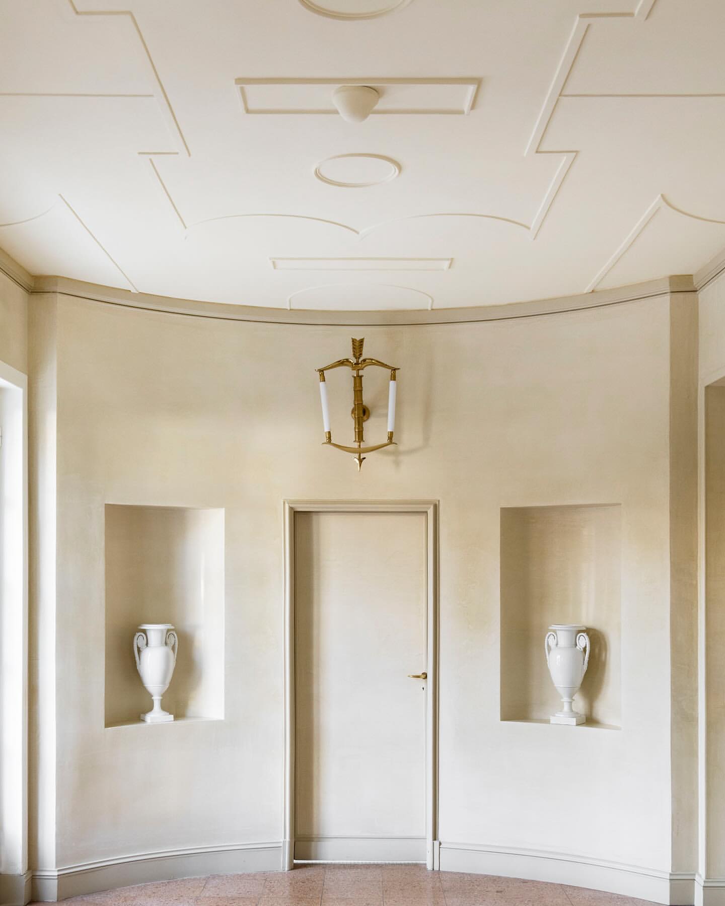 Interior view of a curved hallway featuring white walls with recessed niches holding vases, a door, and an ornate wall sconce.