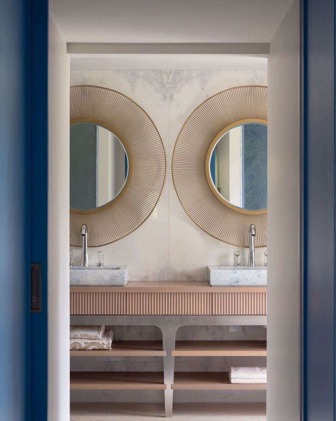 Modern bathroom interior featuring a double vanity with round mirrors, marble countertops, and open shelving for towels, illuminated by soft lighting.