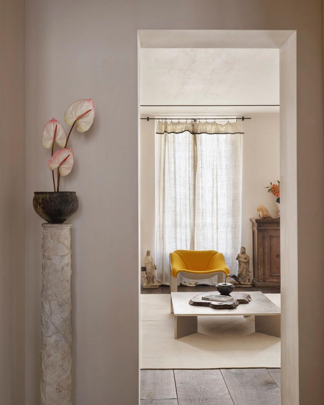 Interior view through an archway featuring a potted plant, a yellow chair, a stone coffee table, and decorative elements in a home.