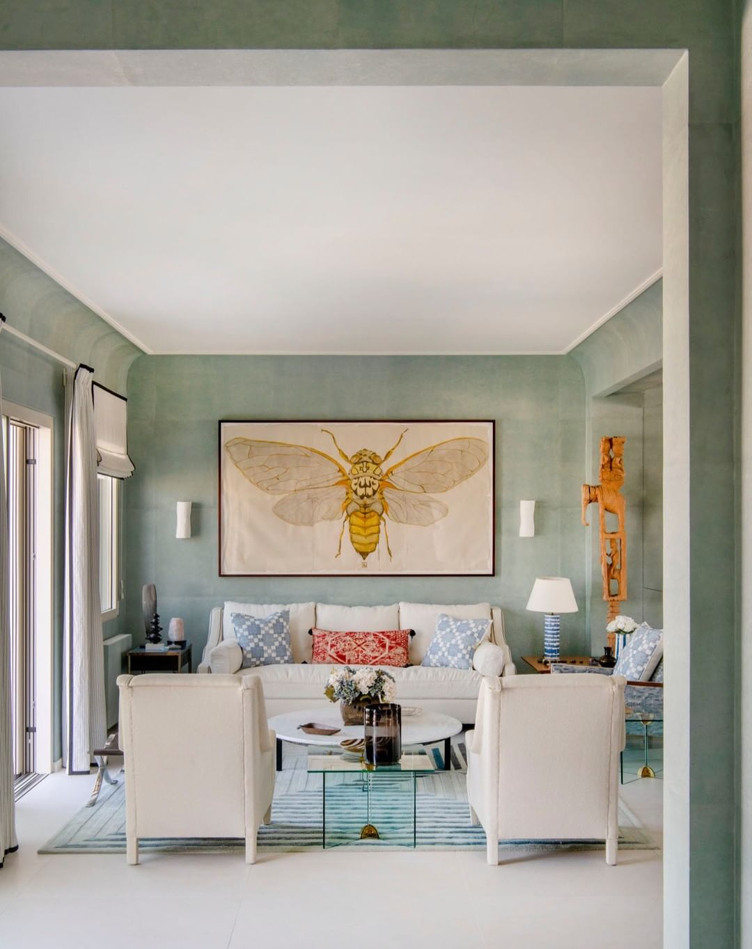 A thoughtfully decorated living room features a neutral sofa with patterned pillows, flanked by white chairs, and an insect print.