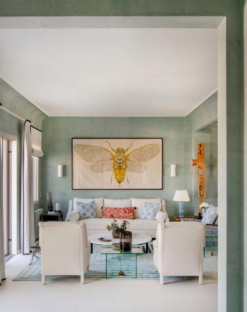 A thoughtfully decorated living room features a neutral sofa with patterned pillows, flanked by white chairs, and an insect print.