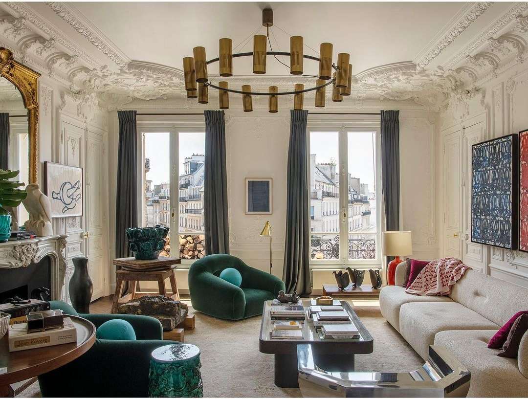 Living room interior with green armchairs, off-white sofa, ornate fireplace, and modern light fixture under decorative ceiling.