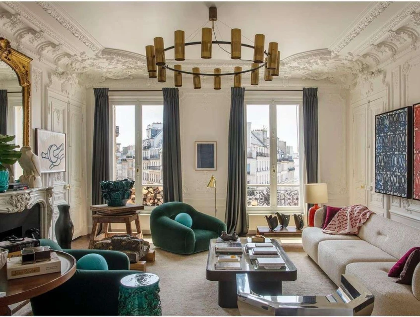 Living room interior with green armchairs, off-white sofa, ornate fireplace, and modern light fixture under decorative ceiling.