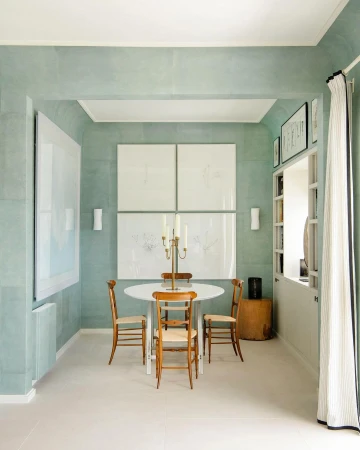 Interior of a dining room featuring a white round table, wooden chairs, and artwork on the wall in a room with light-green walls.