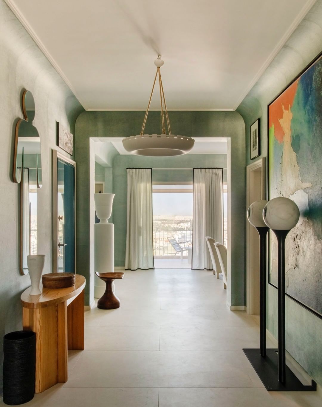View of an interior hallway with modern decor. Console tables, art, and lighting create a stylish residential space. Neutral tones, white floors.
