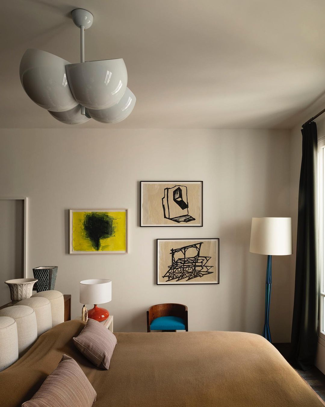 Bedroom features a beige upholstered bed, patterned pillows, three abstract paintings, an off-white lamp, and a unique white chandelier.