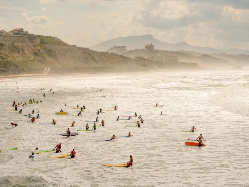 Numerous surfers in the ocean on surfboards with a coastal hillside in the background under a bright, overcast sky.