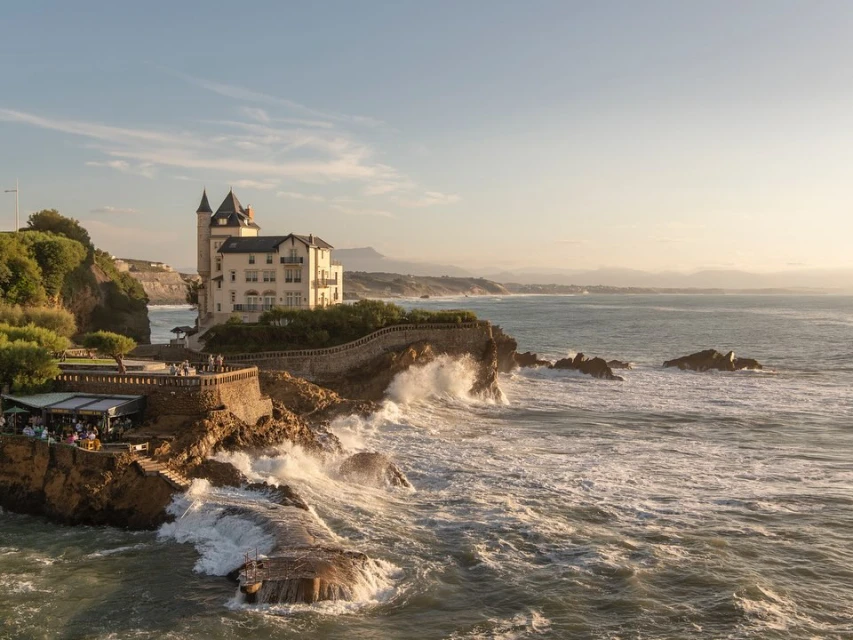 Grand, turreted architecture meets the ocean waves on a rocky coastline, featuring many people on the terrace, under a bright, clear sky.