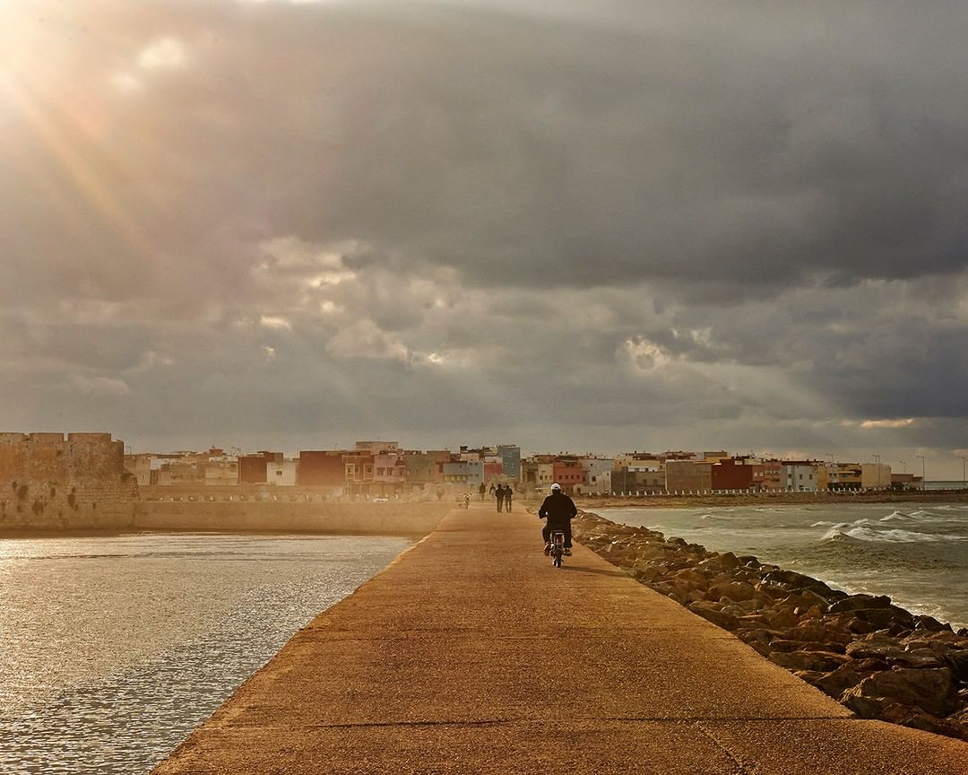 Person rides a motorbike on a coastal road with buildings, rocks, and ocean waves under a cloudy sky and bright sunlight.