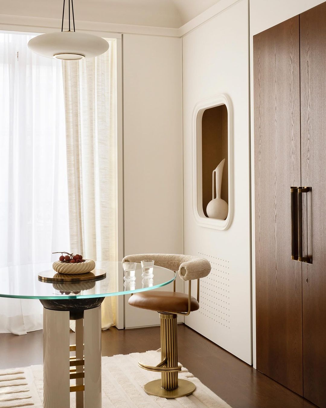 Interior view of dining area with round glass table, modern bar stool, decorative bowl with fruit, and minimalist design elements.