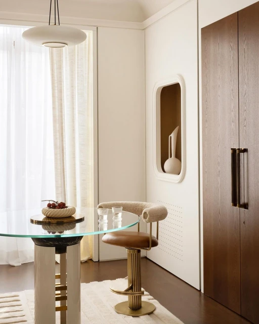 Interior view of dining area with round glass table, modern bar stool, decorative bowl with fruit, and minimalist design elements.