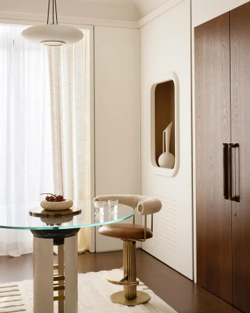 Interior view of dining area with round glass table, modern bar stool, decorative bowl with fruit, and minimalist design elements.