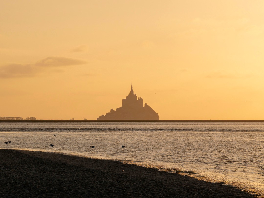Mont Saint-Michel at sunset: A dark silhouette of the iconic abbey stands on a small island surrounded by reflective tidal waters.