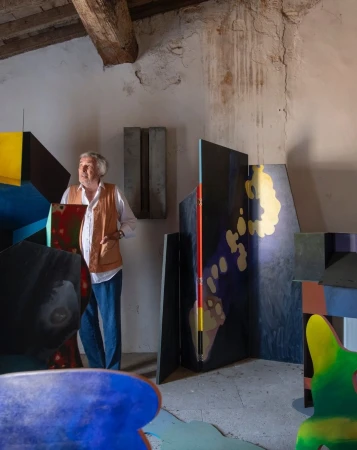 Mature man in a vest stands amid colorful abstract art pieces in a rustic indoor setting with exposed beams and textured walls.