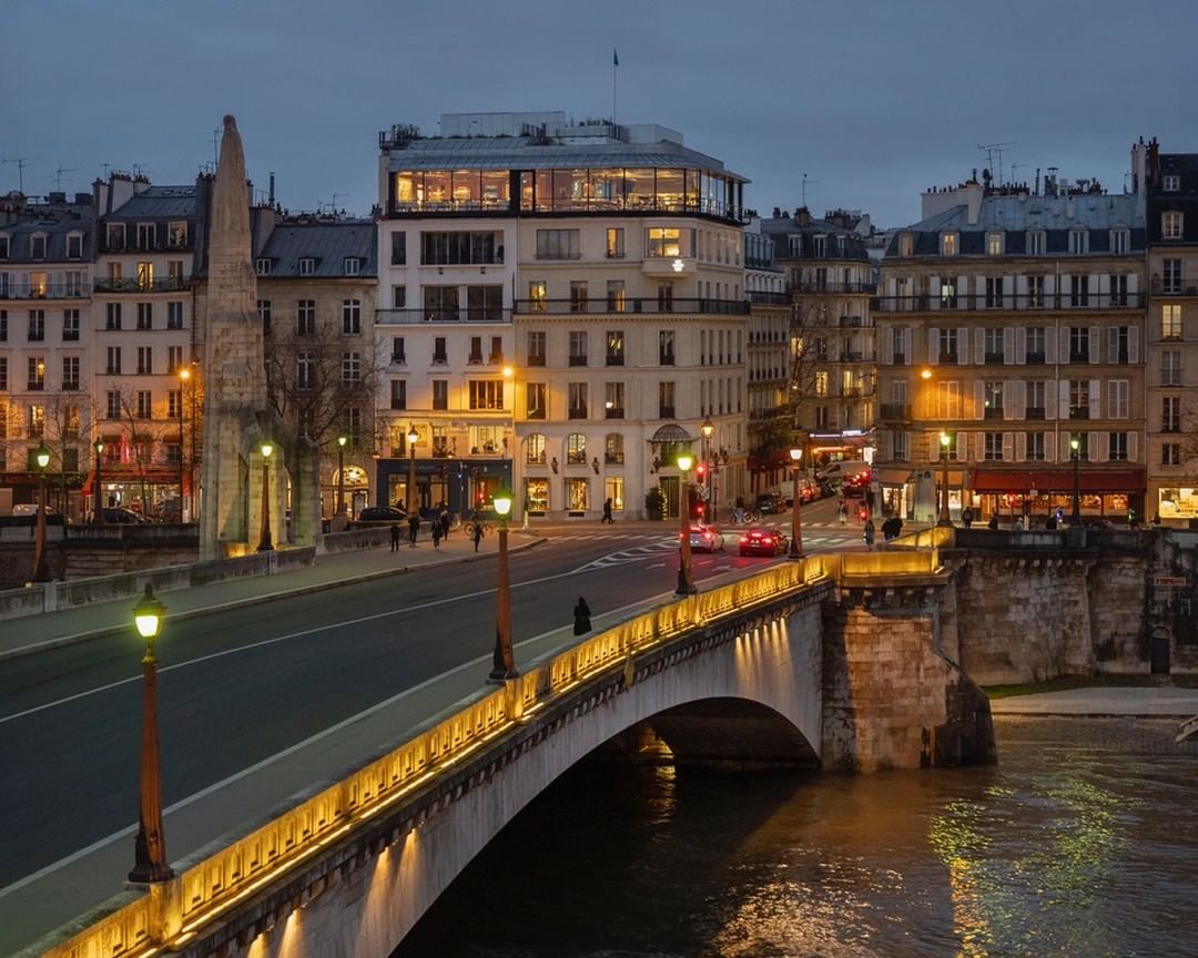 Cityscape of Paris, France at dusk with a bridge over the Seine River, ornate streetlights, and illuminated buildings in the background.