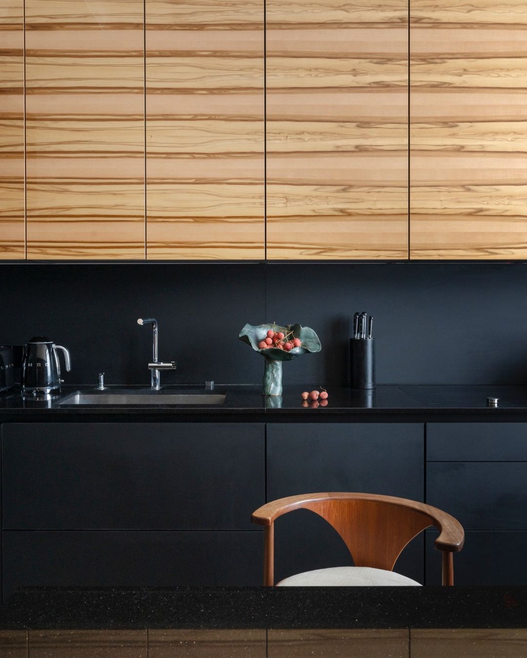 Modern kitchen interior with wooden cabinets, black countertops, stainless steel sink, faucet, and a wooden chair in a home setting.