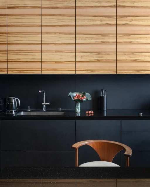 Modern kitchen interior with wooden cabinets, black countertops, stainless steel sink, faucet, and a wooden chair in a home setting.