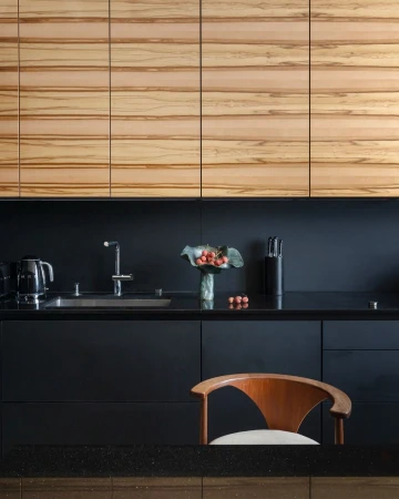 Modern kitchen interior with wooden cabinets, black countertops, stainless steel sink, faucet, and a wooden chair in a home setting.