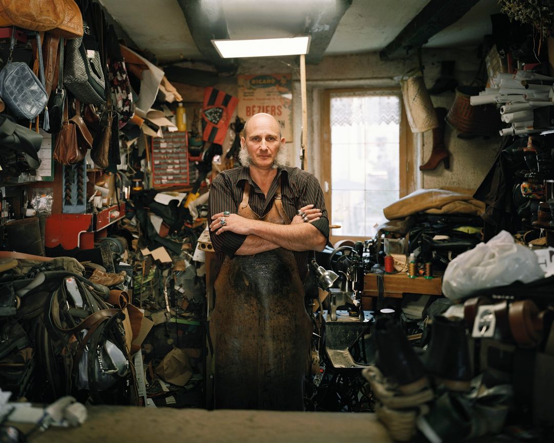 Mature shoemaker stands with crossed arms in workshop surrounded by leather goods, tools, and vintage decor in a medium, eye-level shot.