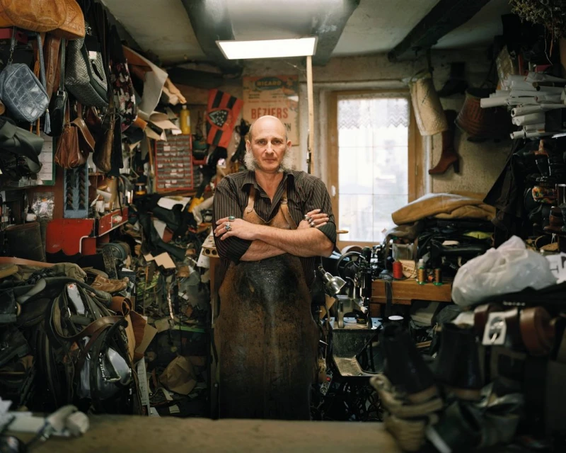 Mature shoemaker stands with crossed arms in workshop surrounded by leather goods, tools, and vintage decor in a medium, eye-level shot.