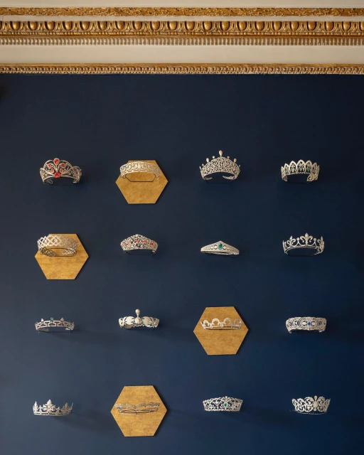 Display of decorative crowns mounted on a dark blue wall with gold hexagonal backings, under an ornate gold and white cornice.