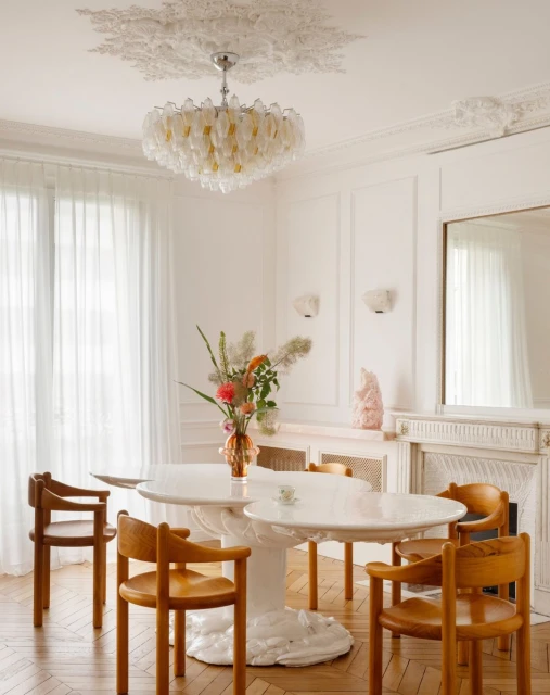 Bright, airy dining room featuring a round marble table, wooden chairs, and a floral centerpiece, illuminated by a modern chandelier.