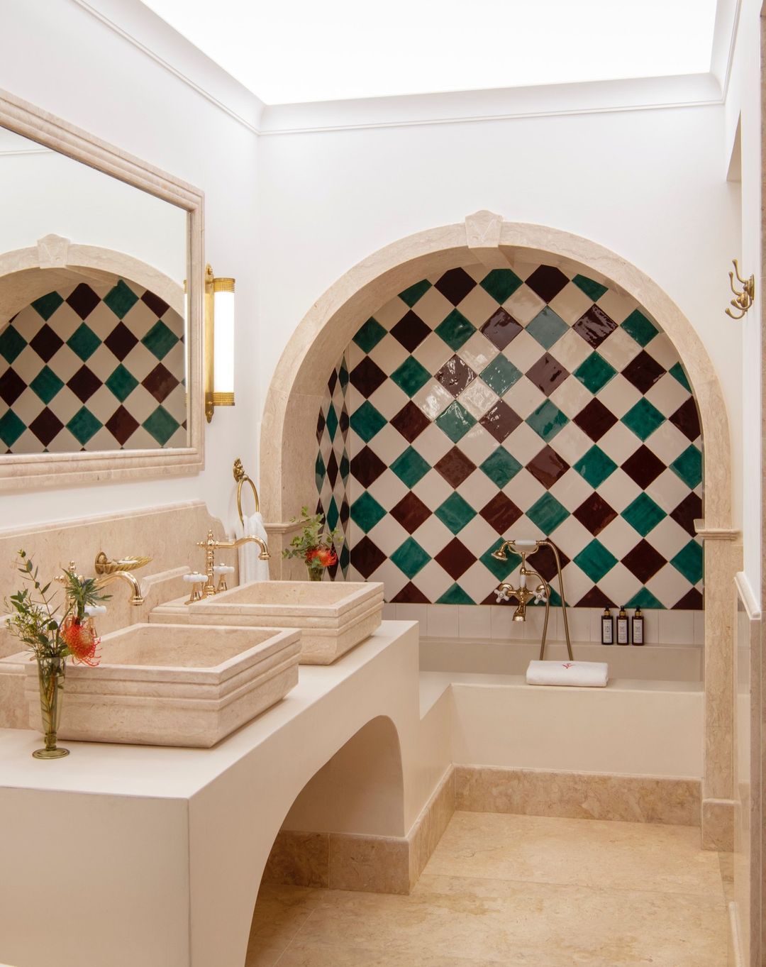 Interior view of a white bathroom with two sinks, gold fixtures, mirror, arched shower tiled in diamond pattern, and marble flooring.