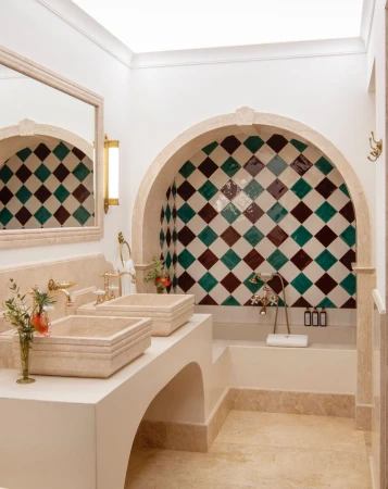Interior view of a white bathroom with two sinks, gold fixtures, mirror, arched shower tiled in diamond pattern, and marble flooring.