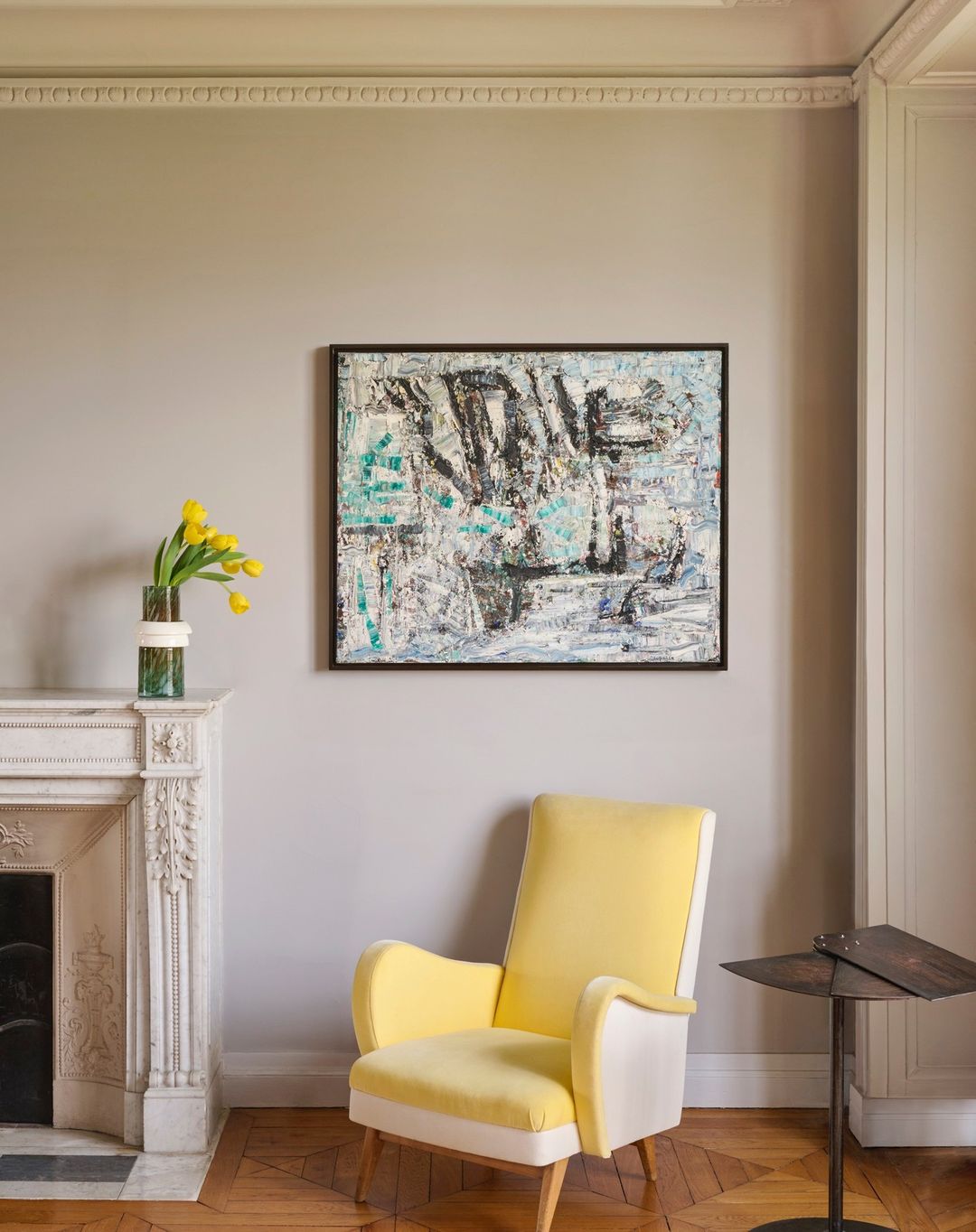 Interior design with abstract painting, yellow armchair, marble fireplace, tulips in a vase, and parquet flooring.