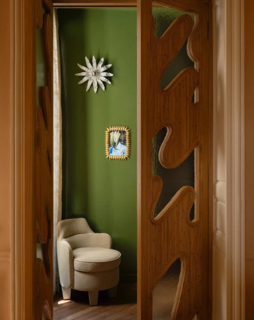 Interior design shot featuring a light beige chair and a decorative carved wooden door in front of an olive green wall with art.