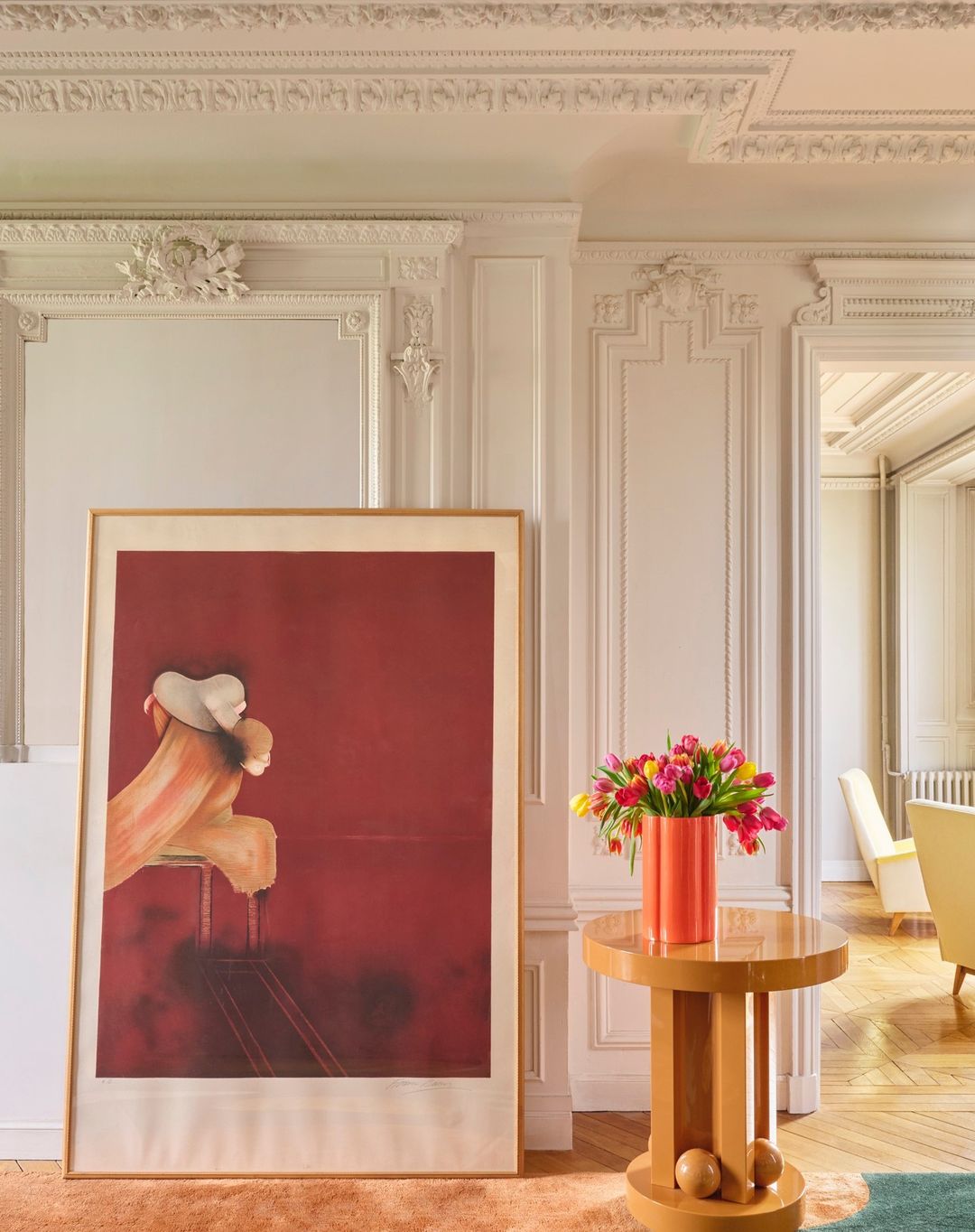 Living room interior featuring a painting, tulip flowers in a vase on a round table, decorative wall molding, and hardwood floors.