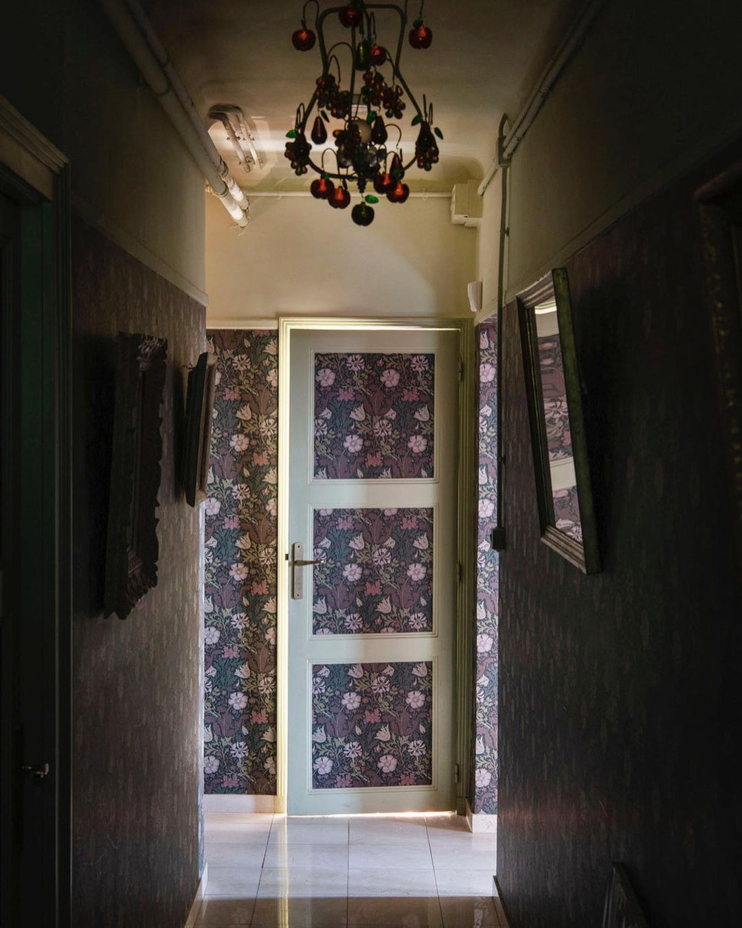 Interior view of a hallway with floral wallpaper, a white door, and a decorative chandelier hanging from the ceiling, warm lighting.
