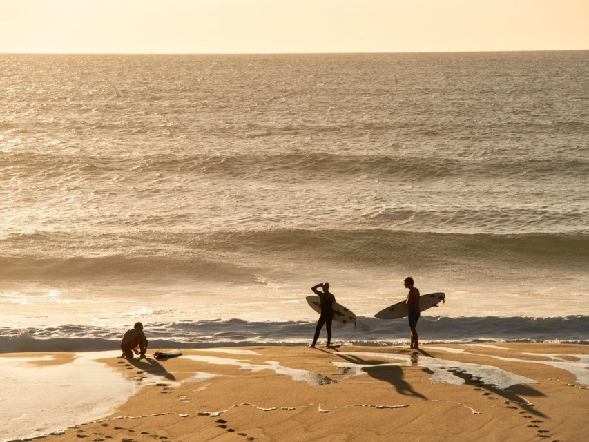Three people with surfboards at sunrise stand on a sandy beach with ocean waves in the background in a wide outdoor landscape shot.