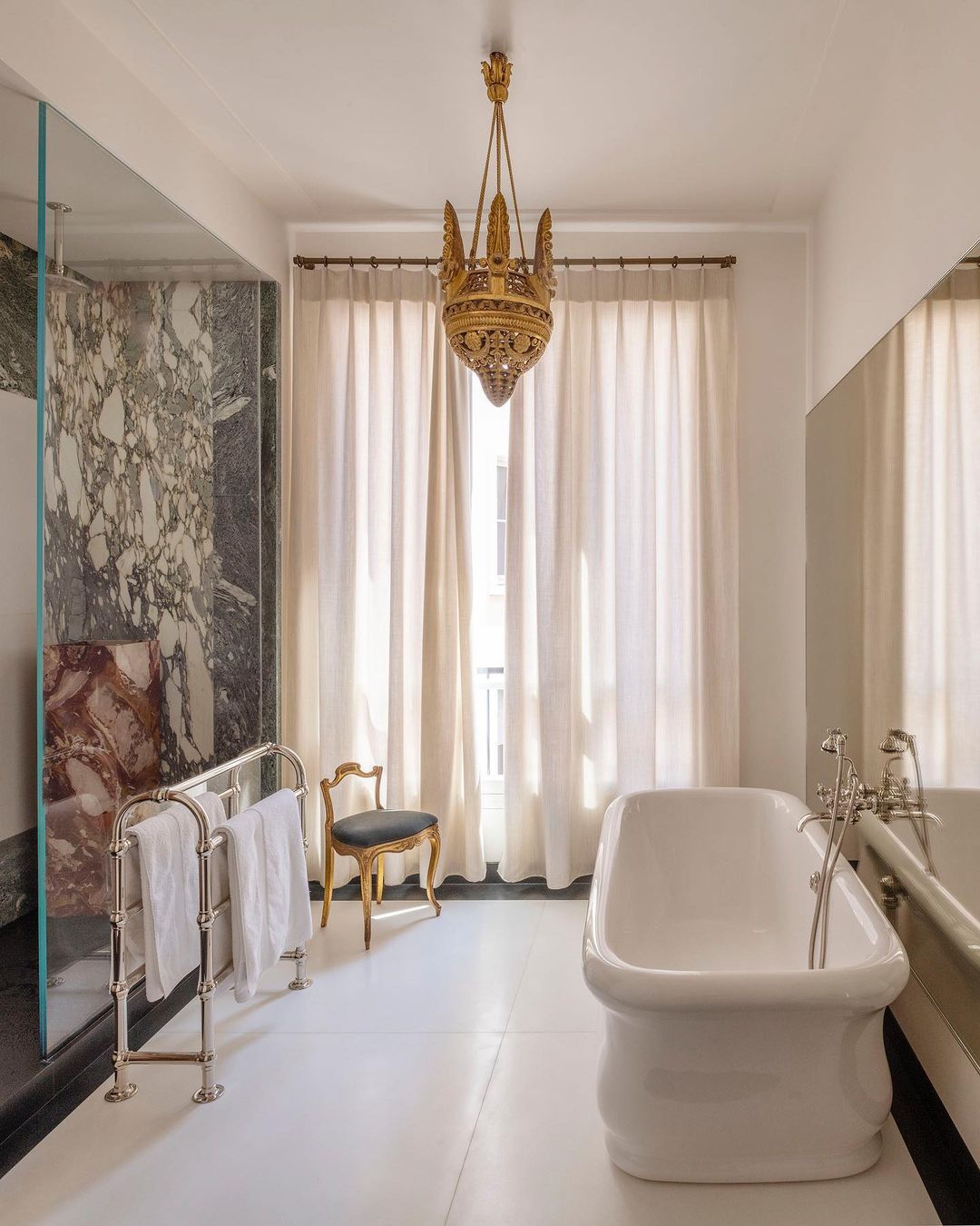 Bright bathroom interior with a white freestanding bathtub, marble wall, ornate lighting fixture, and a small chair by a window.