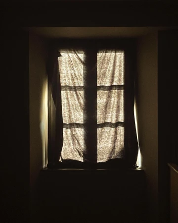 Sunlight streams through a paneled window covered by a neutral-toned curtain in a darkened room, creating a dramatic contrast.