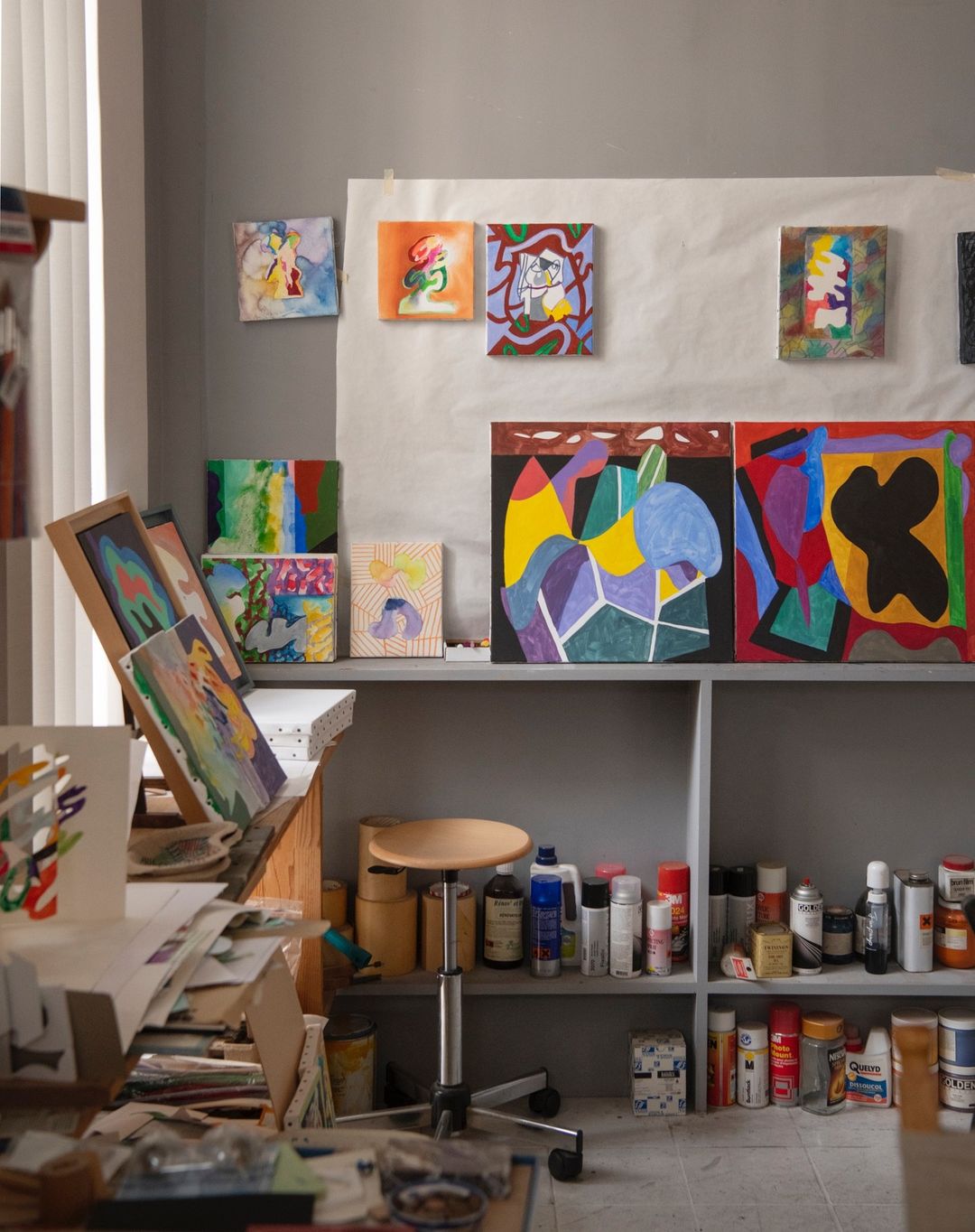A collection of colorful abstract paintings displayed in an artist's studio, featuring various shapes and patterns on canvases and shelves.