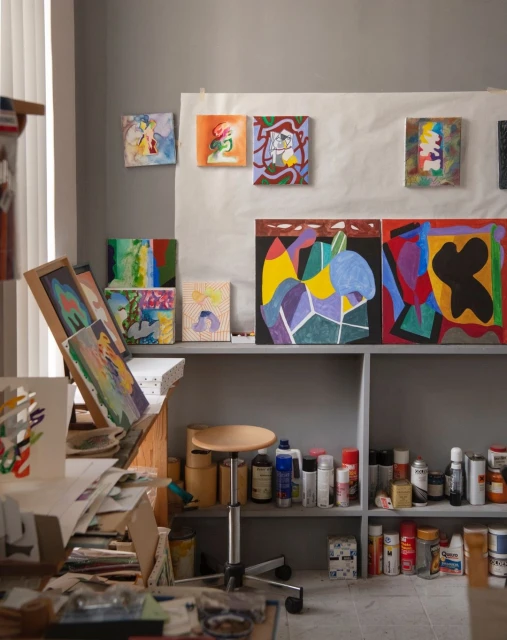 A collection of colorful abstract paintings displayed in an artist's studio, featuring various shapes and patterns on canvases and shelves.
