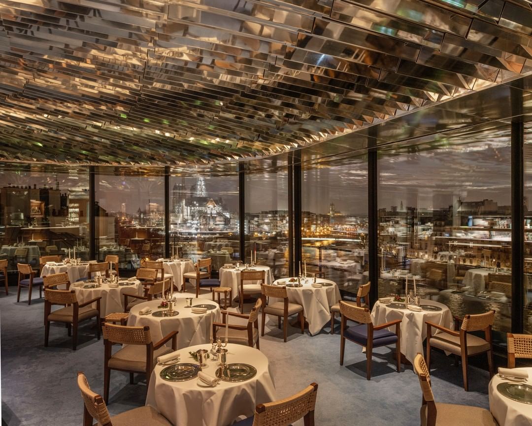 Upscale restaurant interior with tables set for dining, city view through windows, and mirrored ceiling, creating a sophisticated ambiance.