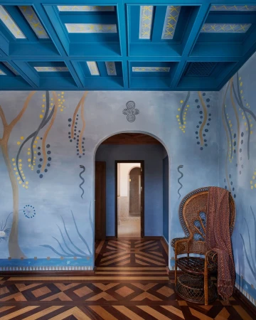 Interior view showcases a wicker chair draped with a brown scarf on a patterned wood floor, set against light blue walls with painted designs.