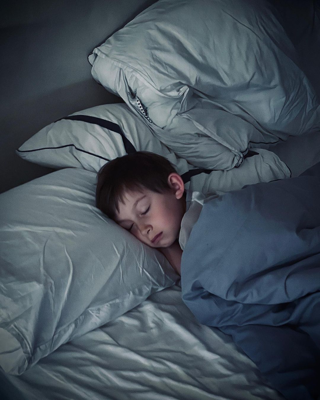 Child sleeps peacefully in bed with white pillows and blue blanket in a dimly lit room at nighttime. Sleeping child in bed.